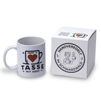 Personalisierte weiße Fototasse von Tasse mit Herz neben einem edlen weißen Geschenkkarton mit dem runden Siegel „Handveredelt im Schwarzwald“.